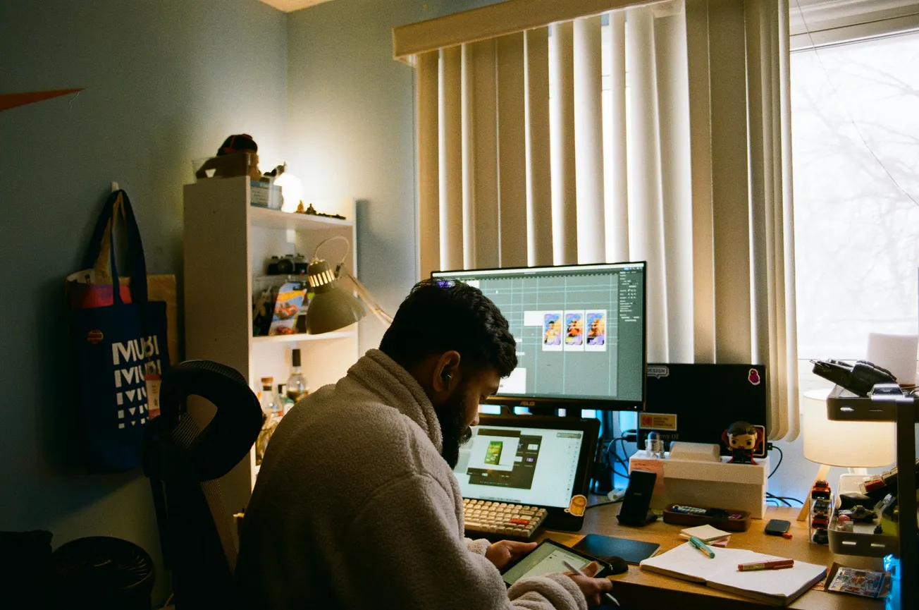 A person in a cozy sweater works at a cluttered desk with dual monitors displaying digital art. A window with blinds casts soft light, creating a focused atmosphere.