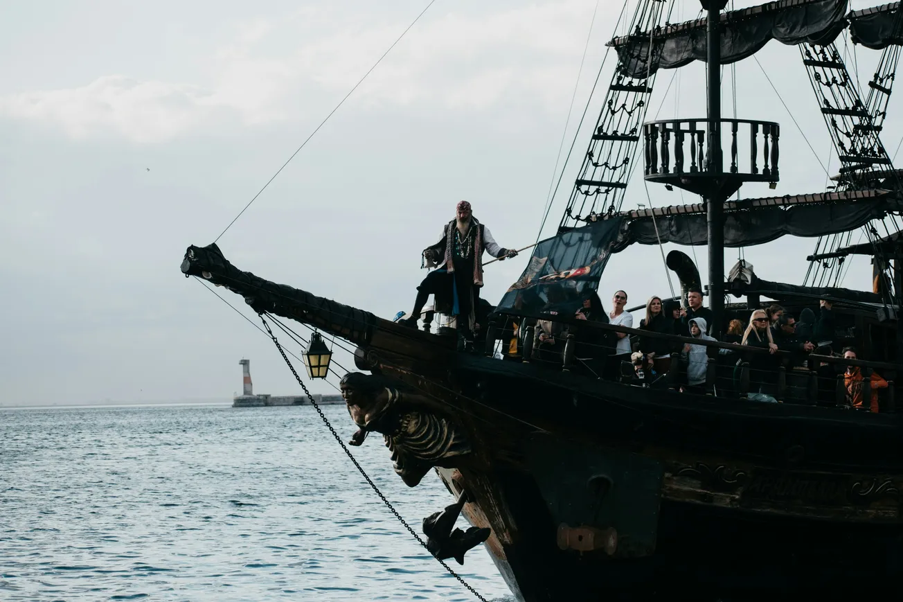 A pirate-themed ship sails with a costumed man on the bow, enthusiastically gesturing. Passengers watch, creating an adventurous and lively scene.