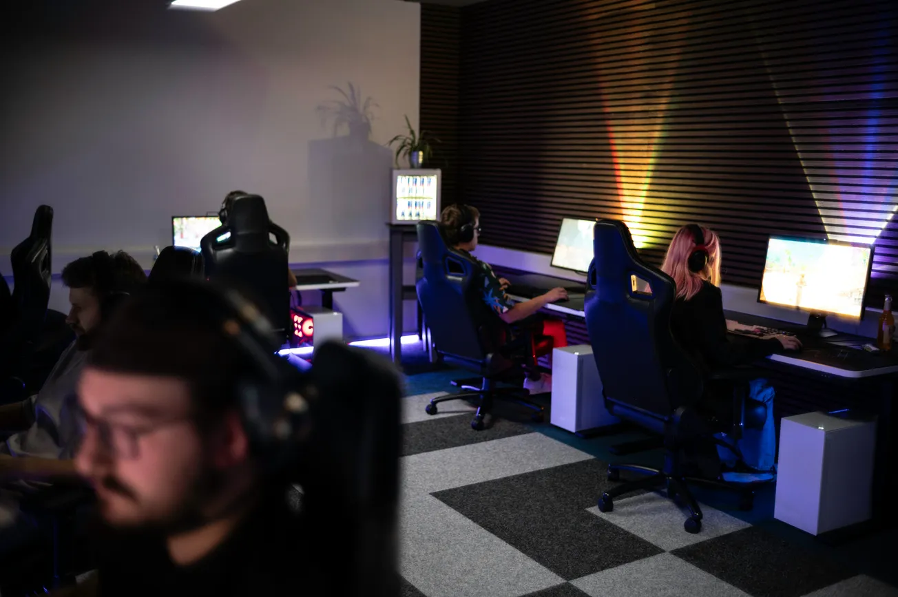 A dimly lit gaming room with four people focused on computer screens. Colorful lights reflect on the walls, creating an immersive atmosphere.