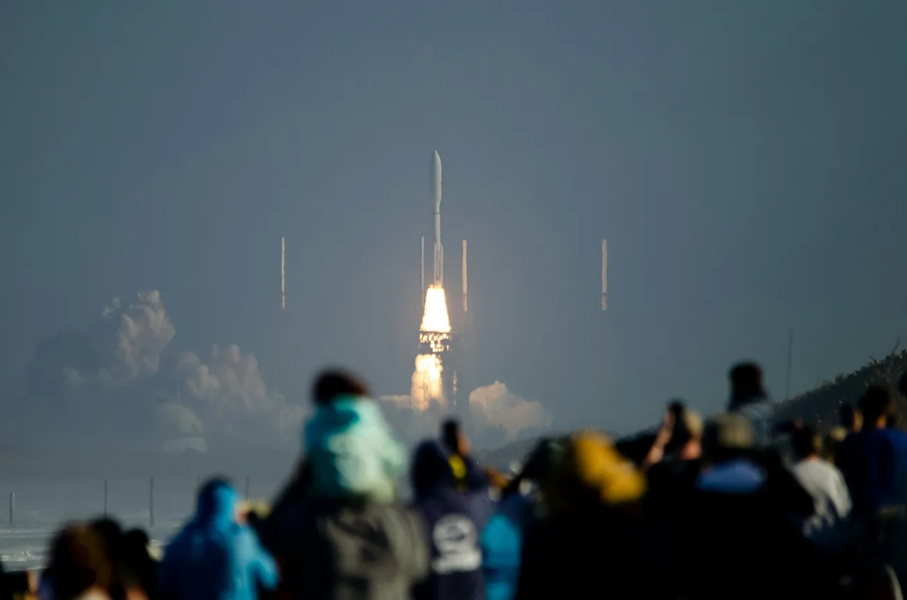 A rocket launches into the sky, leaving behind a trail of fire and smoke. A crowd of people, blurred in the foreground, watches in awe.