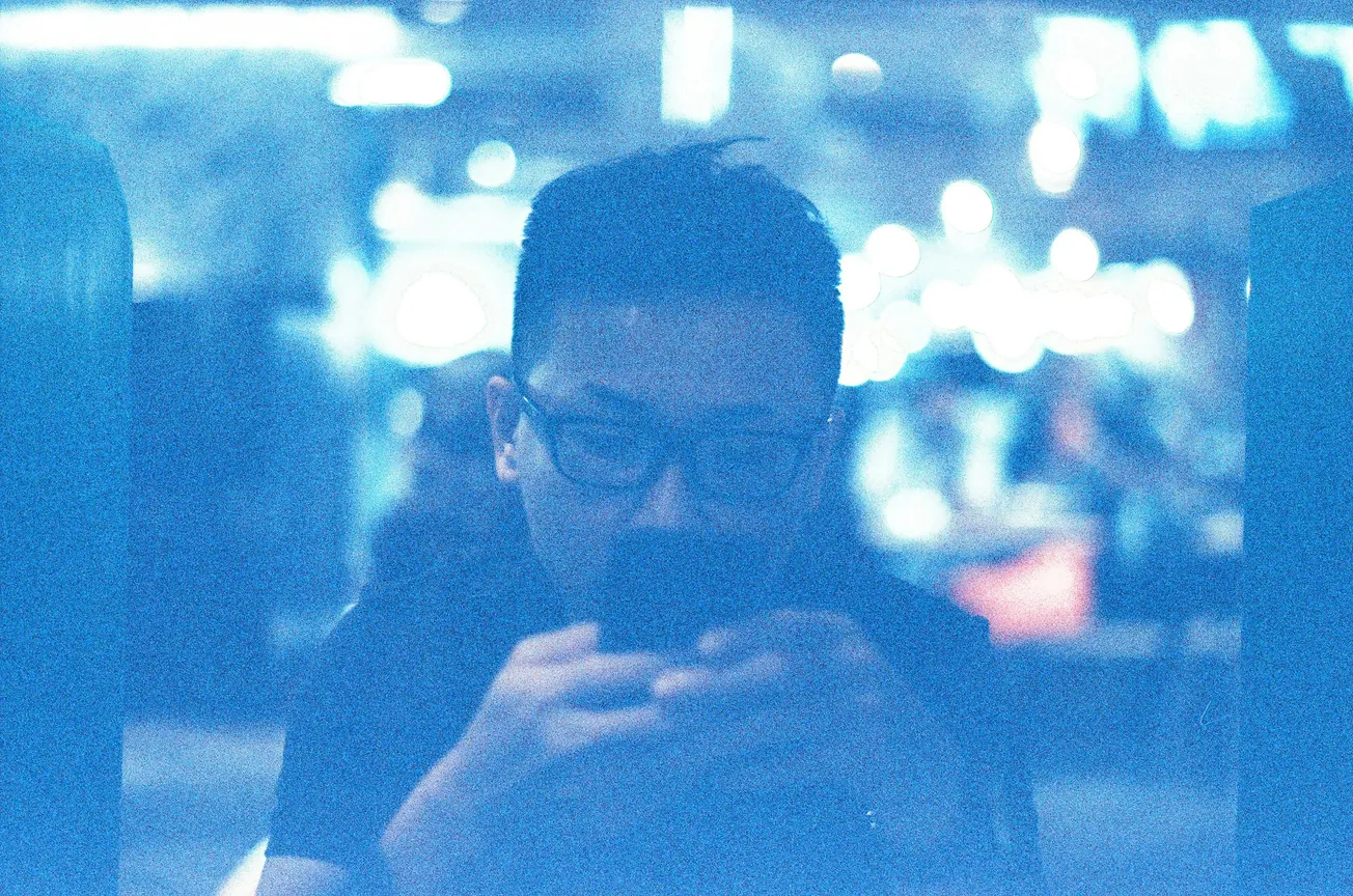 A man with glasses is focused on his phone in a dimly lit, blurry setting with an intense blue tint. The background displays bokeh lights, creating a dreamy ambiance.