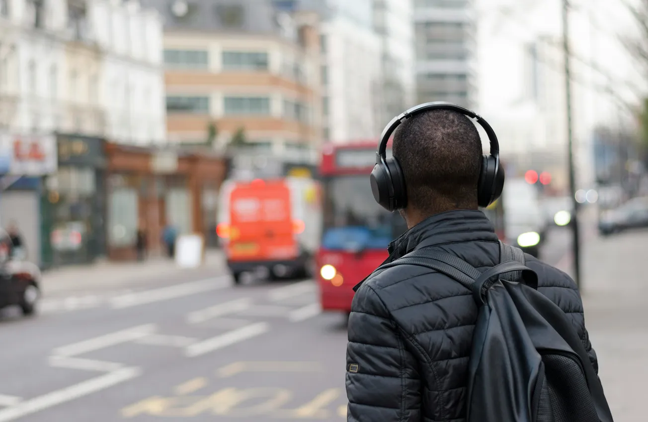 A person in a dark jacket and headphones stands on a city street with traffic, including a red bus. The scene conveys urban life and focus.