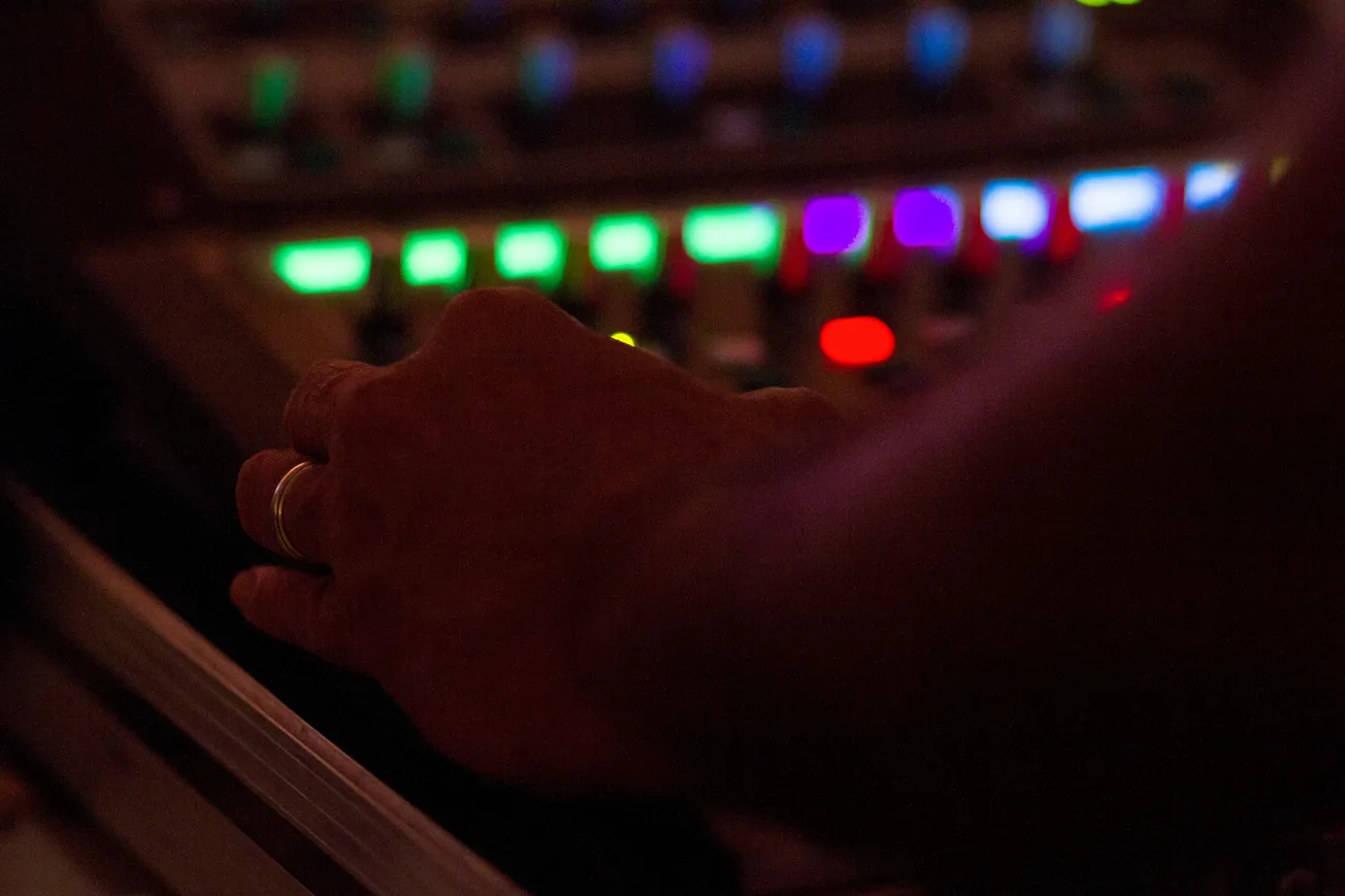 A hand adjusts controls on an audio mixing board, surrounded by colorful, softly glowing lights. The scene is dimly lit, conveying focus and concentration.