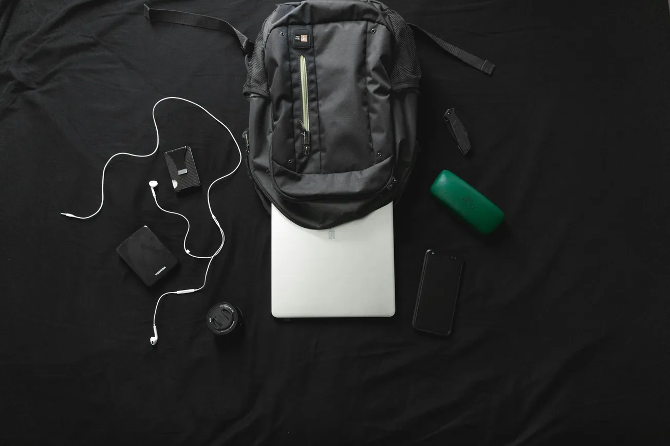 A backpack, laptop, phone, earbuds, wallet, portable speaker, watch, and a camera lens are neatly arranged on a black background.