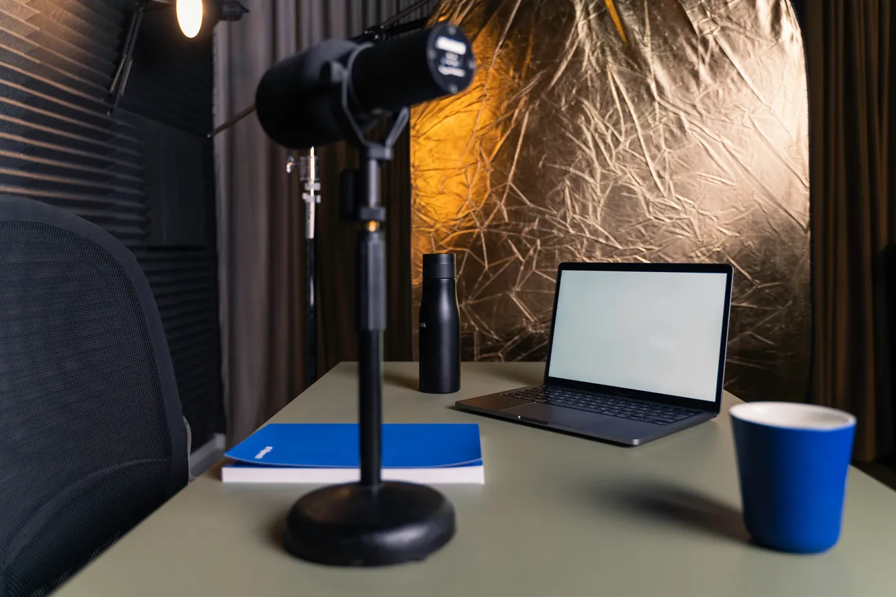 A podcast studio setup with a microphone, laptop, blue notebook, and coffee cup on a desk. Soft lighting and soundproofed walls create a cozy atmosphere.