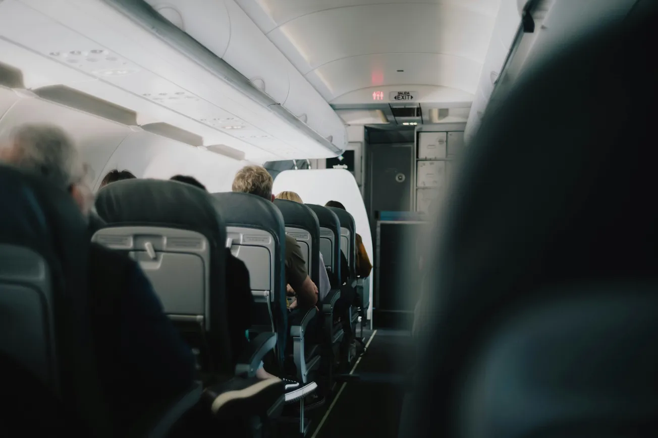 Interior of an airplane cabin with rows of occupied seats. Passengers are seated facing forward, and the dimly lit aisle leads to the rear exit door.