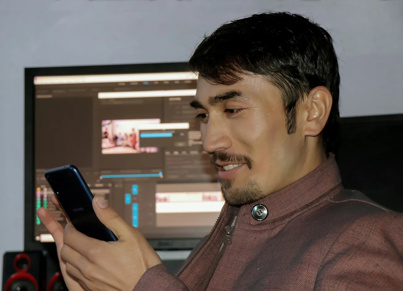 A man in a maroon jacket smiles while looking at his phone. Behind him, a computer screen displays video editing software. He appears focused and content.