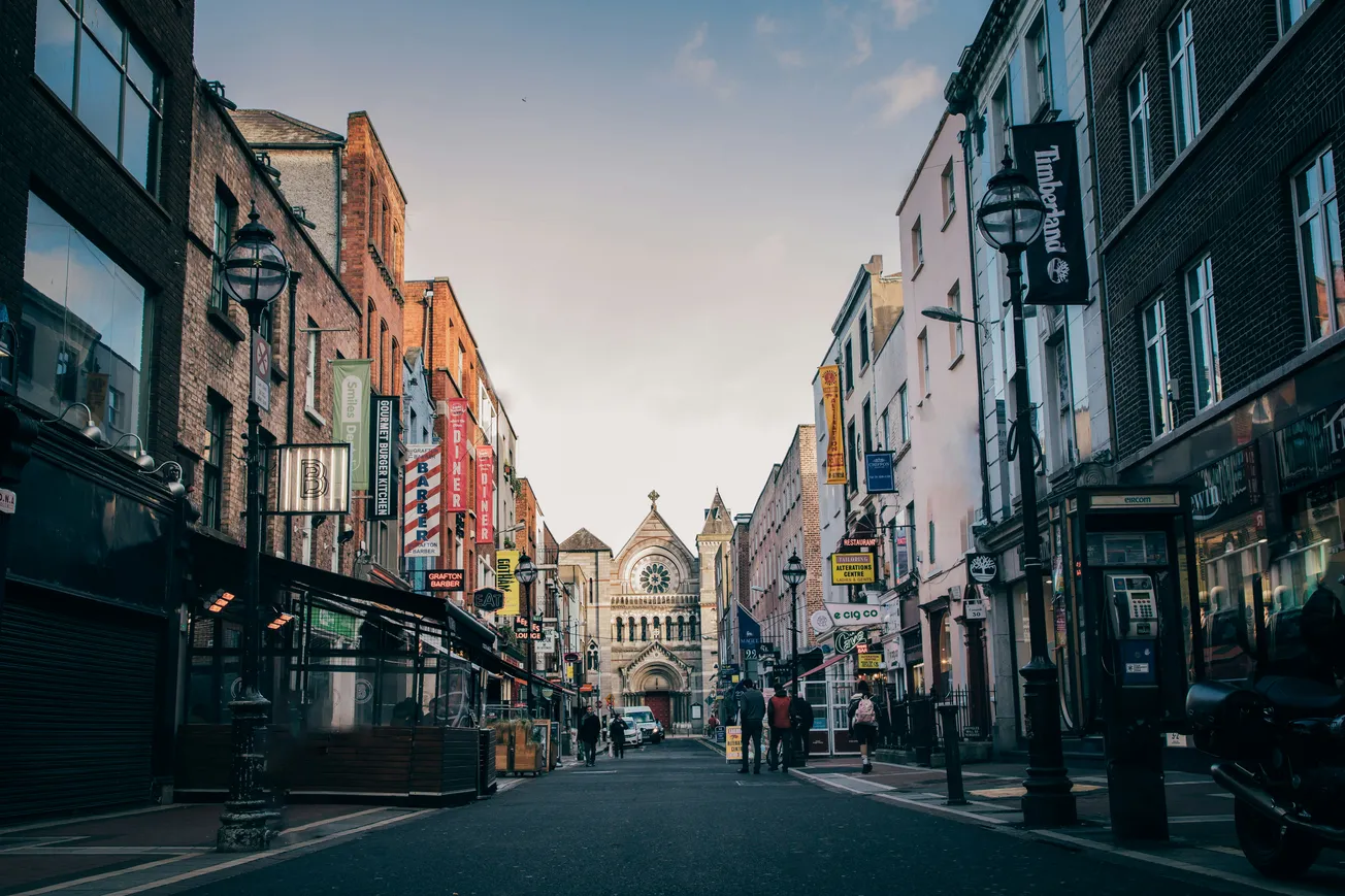 A bustling street lined with shops and colorful signs leads to an ornate church with a large rose window at dusk, creating a lively yet peaceful urban scene.
