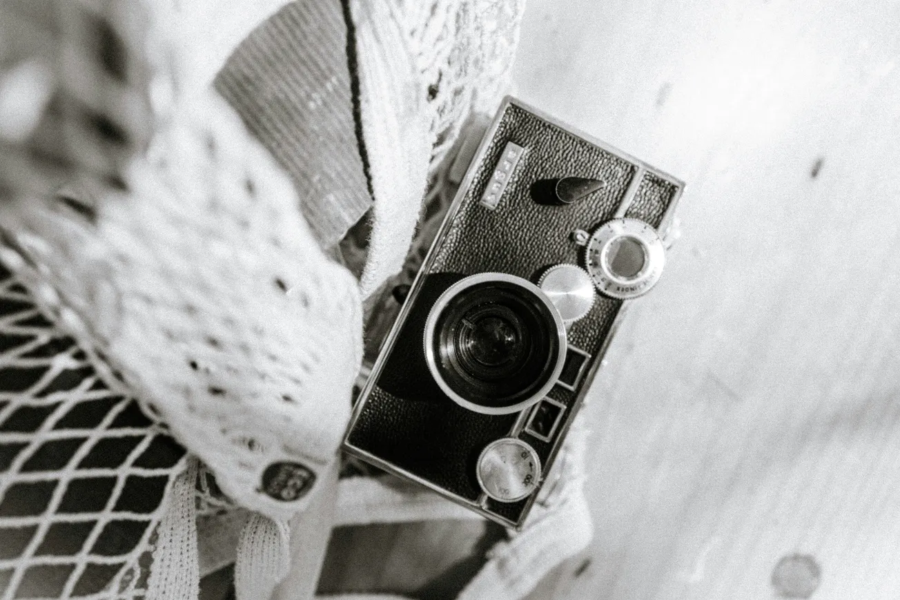 Black and white image of a vintage camera resting on a textured fabric. The camera's large lens and dials evoke a nostalgic, classic feel.