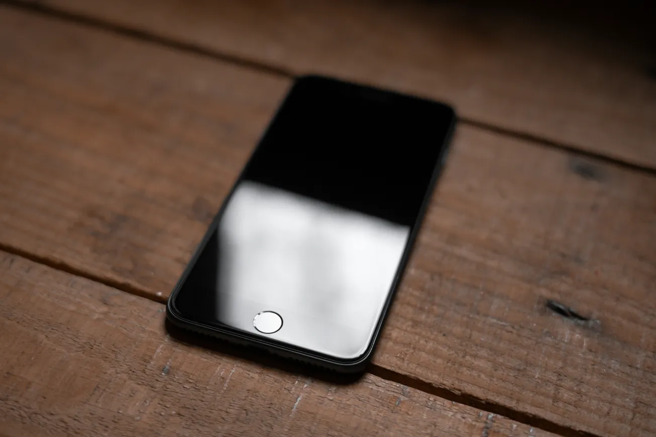 A smartphone with a reflective screen lies on a rustic wooden surface. The lighting creates a soft shadow, and the phone's sleek design is emphasized.