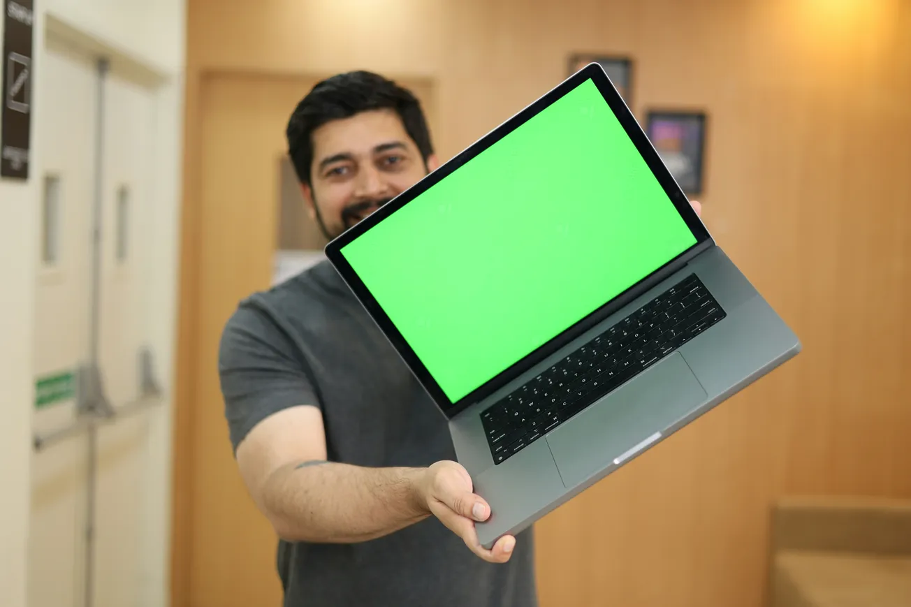 Man holding a laptop with a bright green screen, standing in a room with beige walls. The setting is casual and informal, conveying a sense of approachability.
