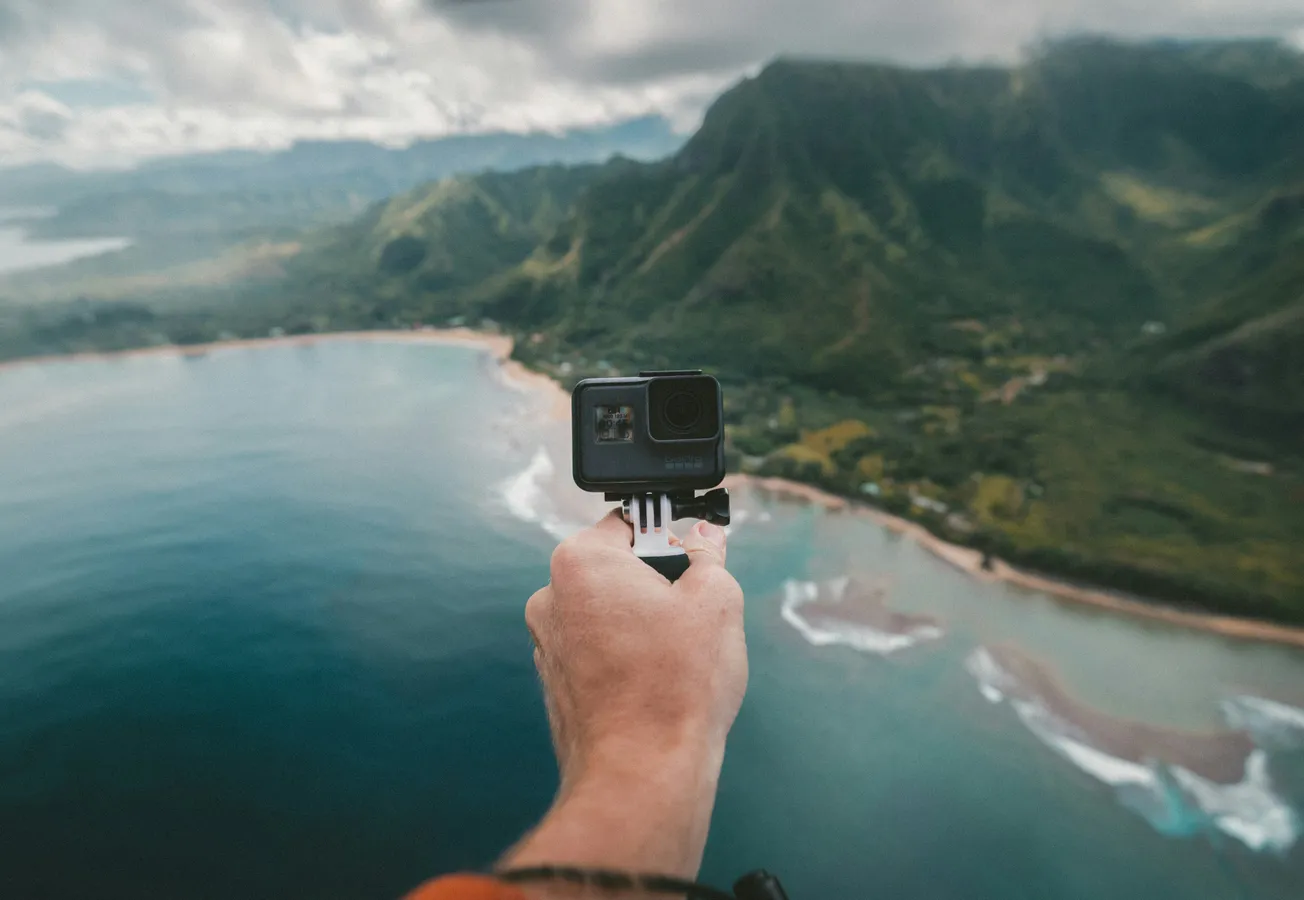 A hand holds a GoPro against a backdrop of lush green mountains and a vast ocean. The image conveys adventure and a sense of freedom.