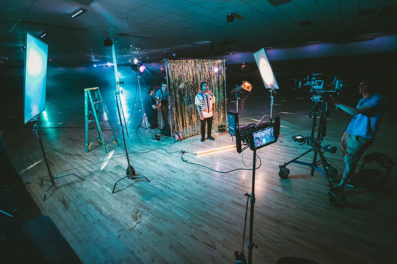 A photo shoot setup in a dimly lit studio features a person standing in front of a gold tinsel backdrop. Bright studio lights and cameras surround them.