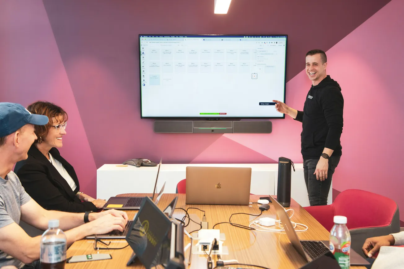 A man presents at a screen displaying charts and data in a modern office. Three colleagues, seated at a table with laptops, watch attentively, smiling.