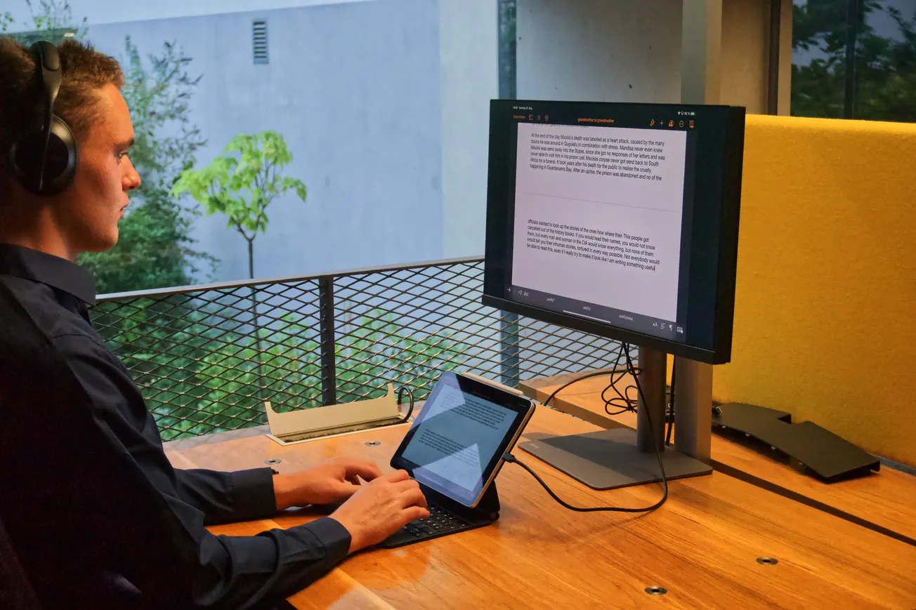 A person wearing headphones works at a wooden desk, typing on a tablet with content mirrored onto a larger screen. The setting is modern and serene.