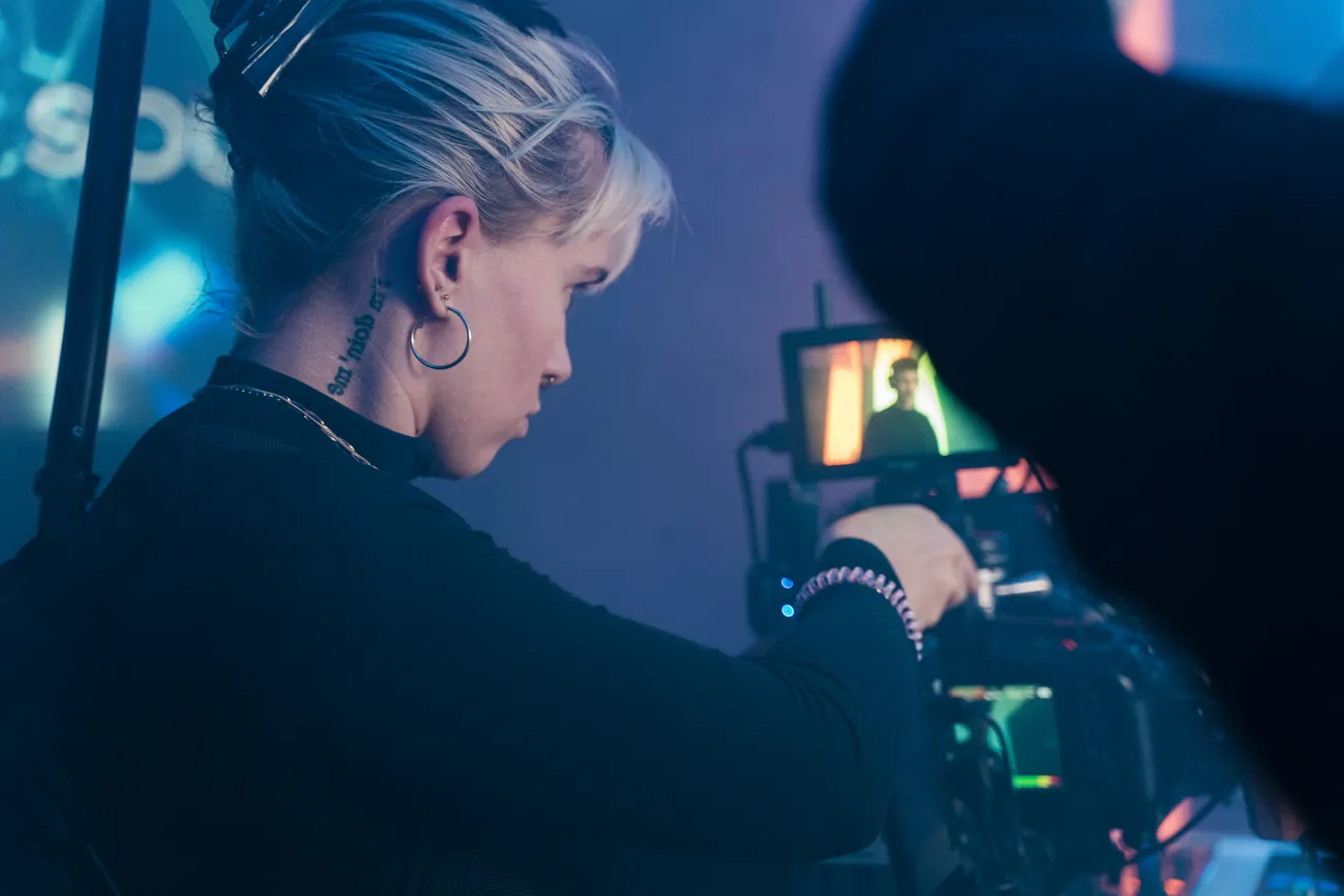 A focused woman with short blonde hair operates a film camera, adjusting settings. A monitor shows a lit scene. The atmosphere is professional and creative.