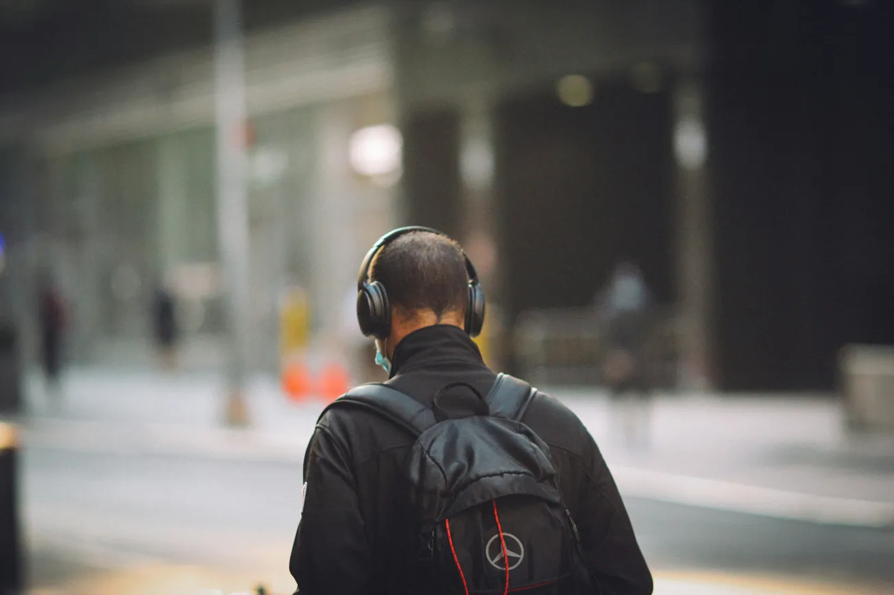 A person with short hair, wearing headphones and a backpack, walks down a city street. The scene is blurred, creating a calm, urban atmosphere.