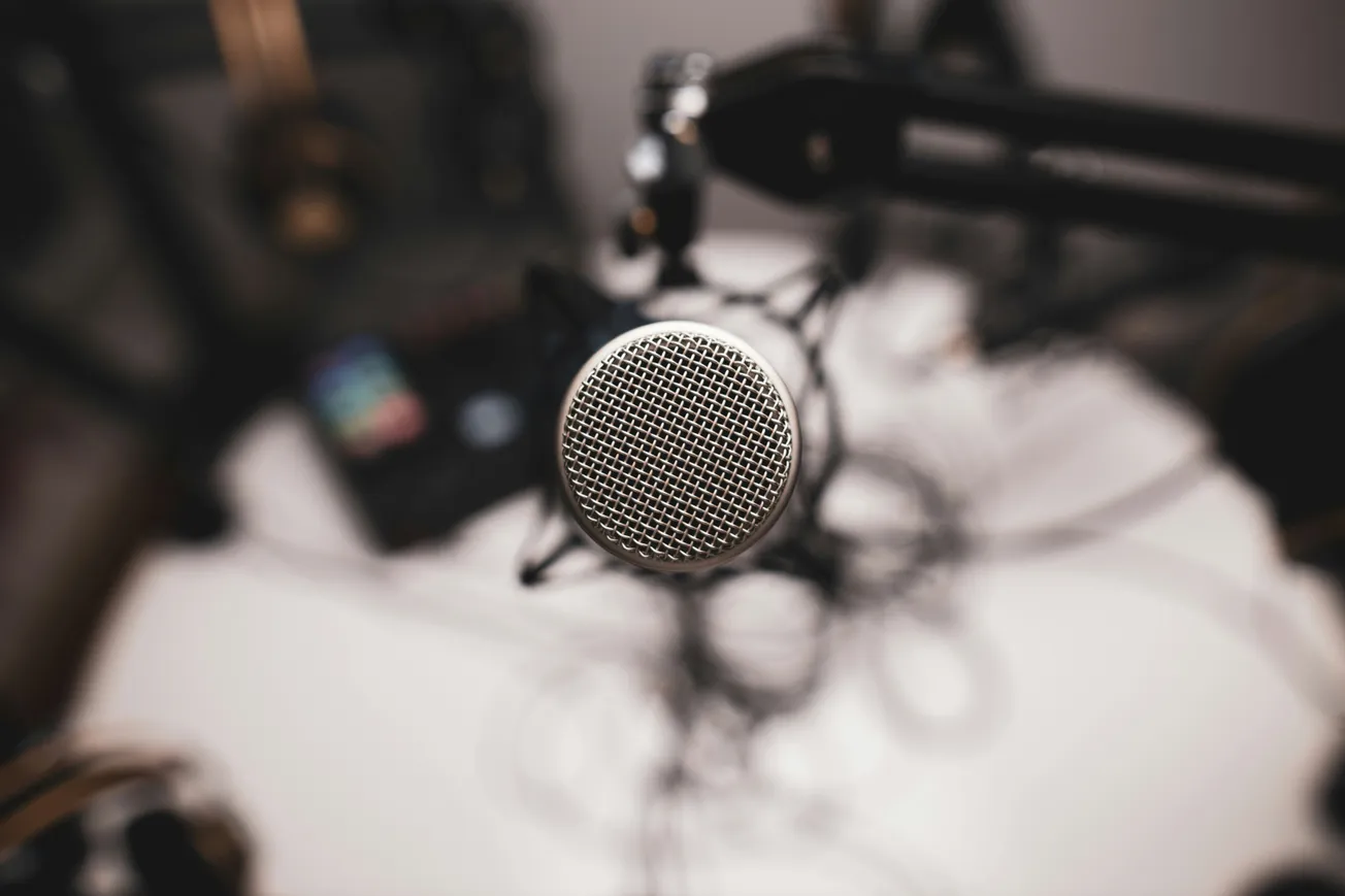 Close-up of a silver microphone on a boom arm, set against a blurred background with audio equipment, suggesting a podcast or recording studio vibe.
