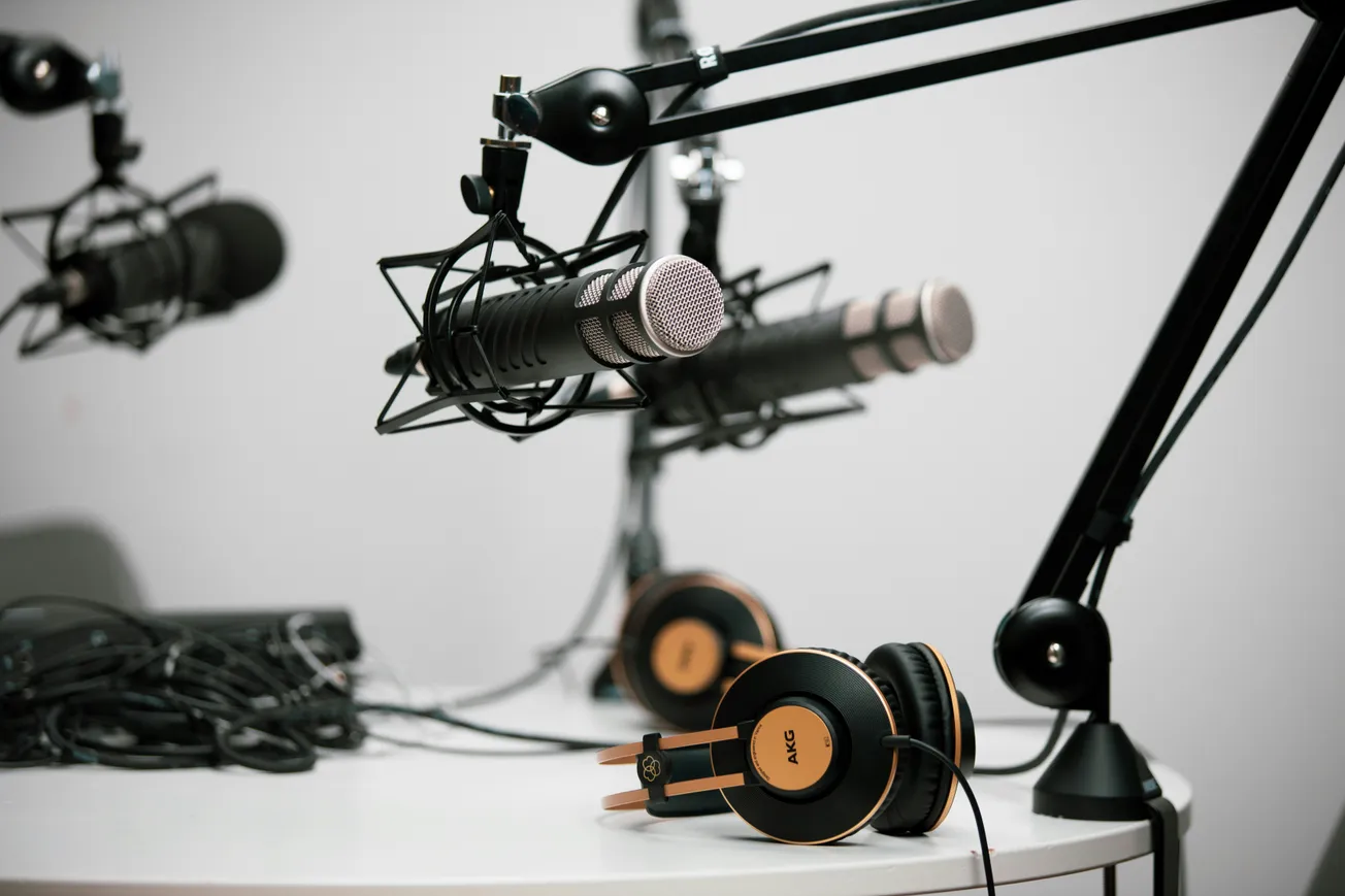 Close-up of a podcast studio setup with two black microphones on adjustable arms and a pair of black and orange headphones on a white table.