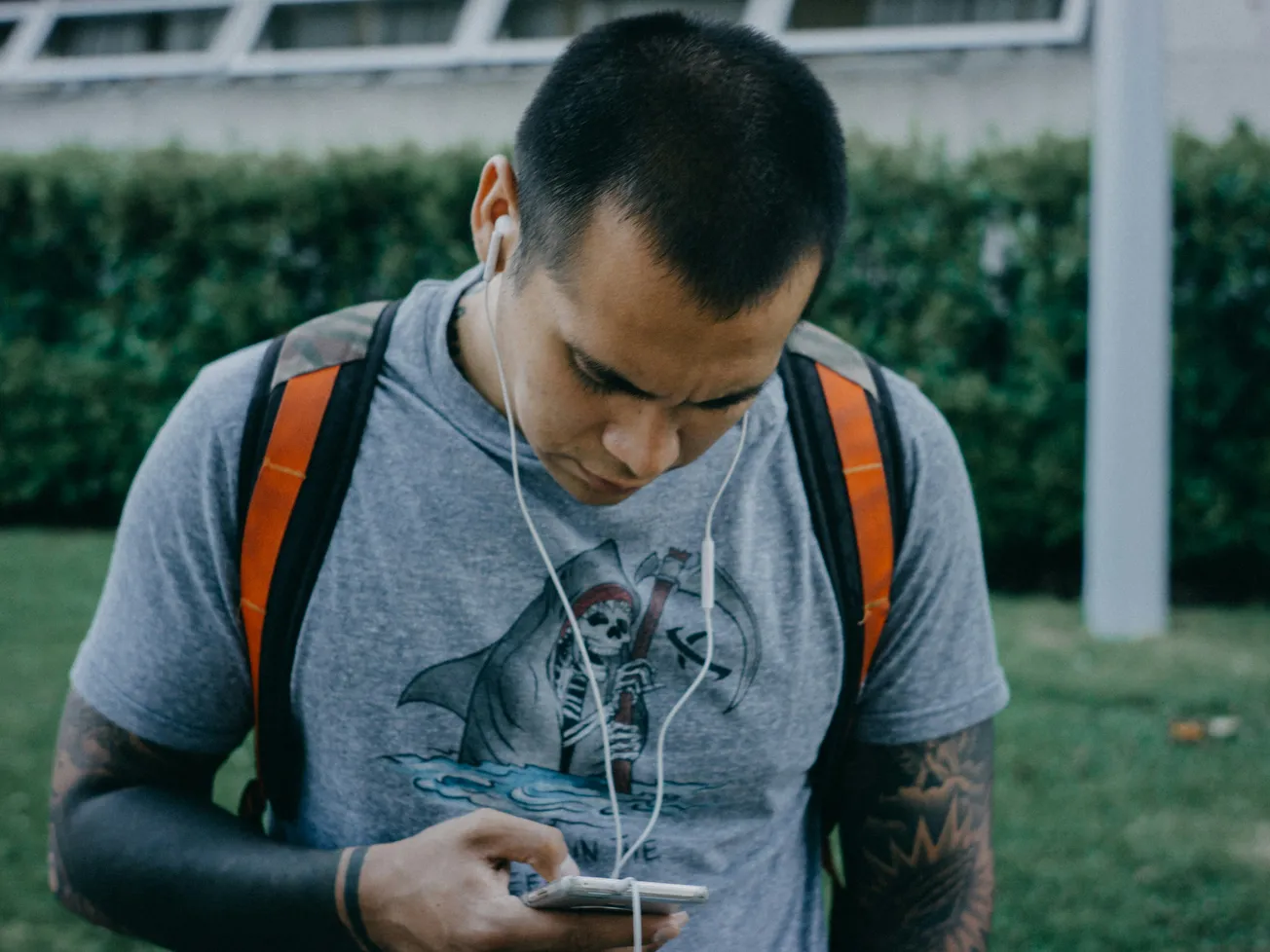 A man wearing a gray t-shirt with a large graphic listens to earphones while looking at his smartphone. He has tattoos, a backpack, and stands outdoors.