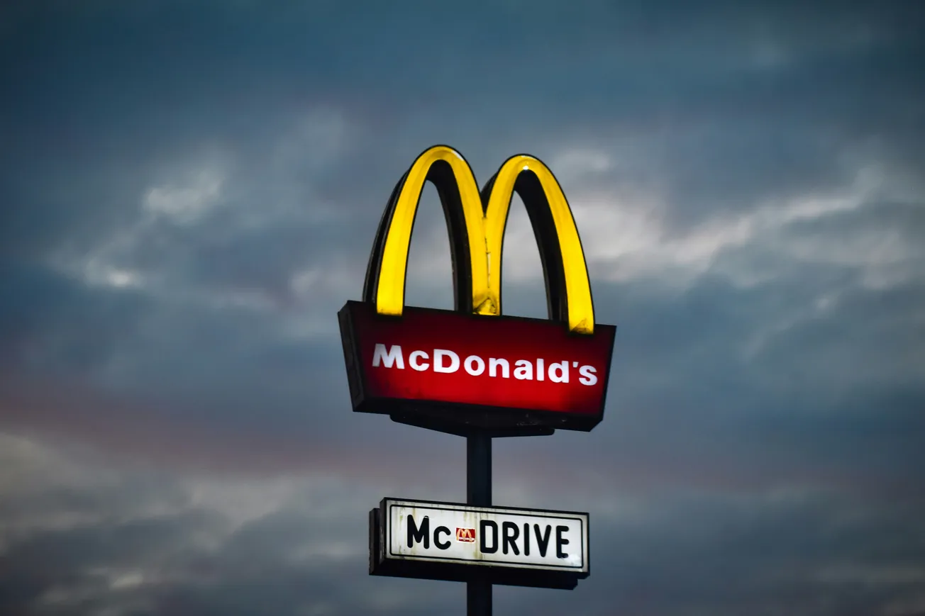 A lit McDonald's sign with golden arches and "McDrive" text is set against a dramatic evening sky with dark clouds, conveying a moody atmosphere.
