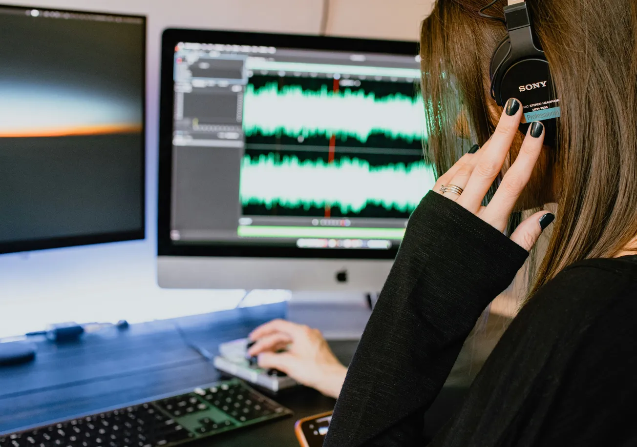 A woman with headphones edits audio on dual monitors displaying sound waves. The setting is a modern workstation, conveying focus and tech-savvy.