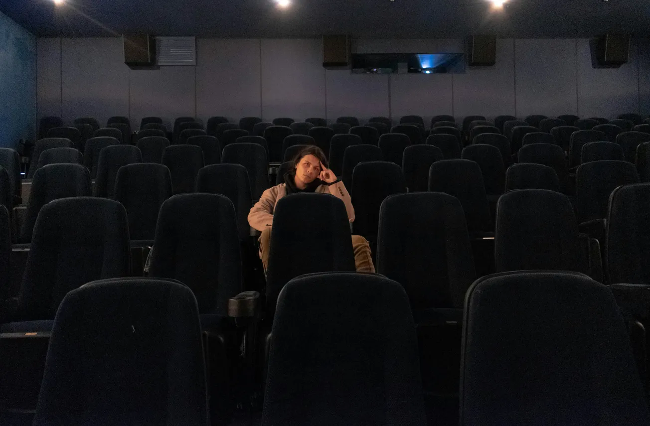 Person sitting alone in a dimly lit, almost empty theater. Rows of dark seats surround them, creating a contemplative and solitary atmosphere.