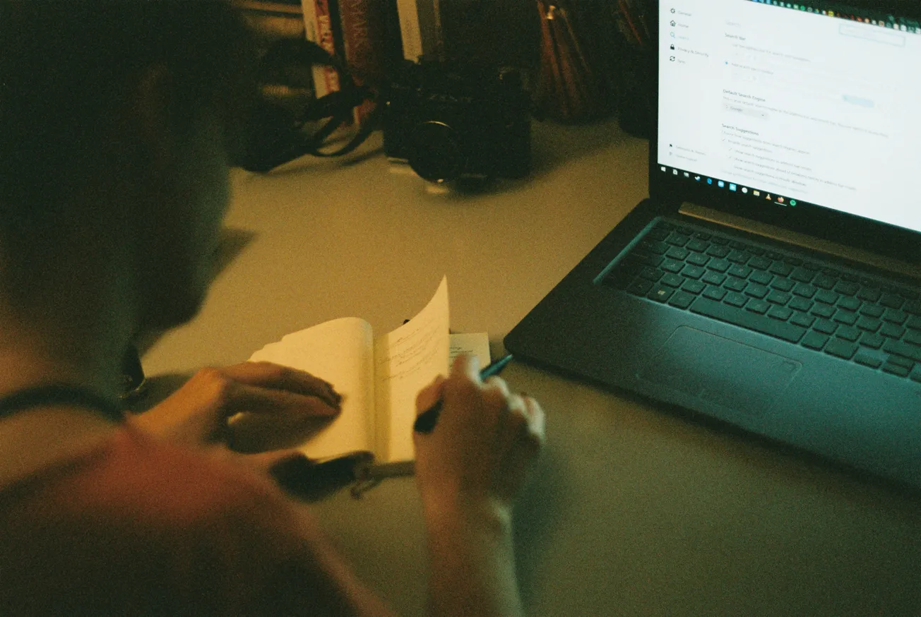 A person writes in a notebook next to an open laptop on a desk. The scene is dimly lit, creating an atmosphere of focus and concentration.