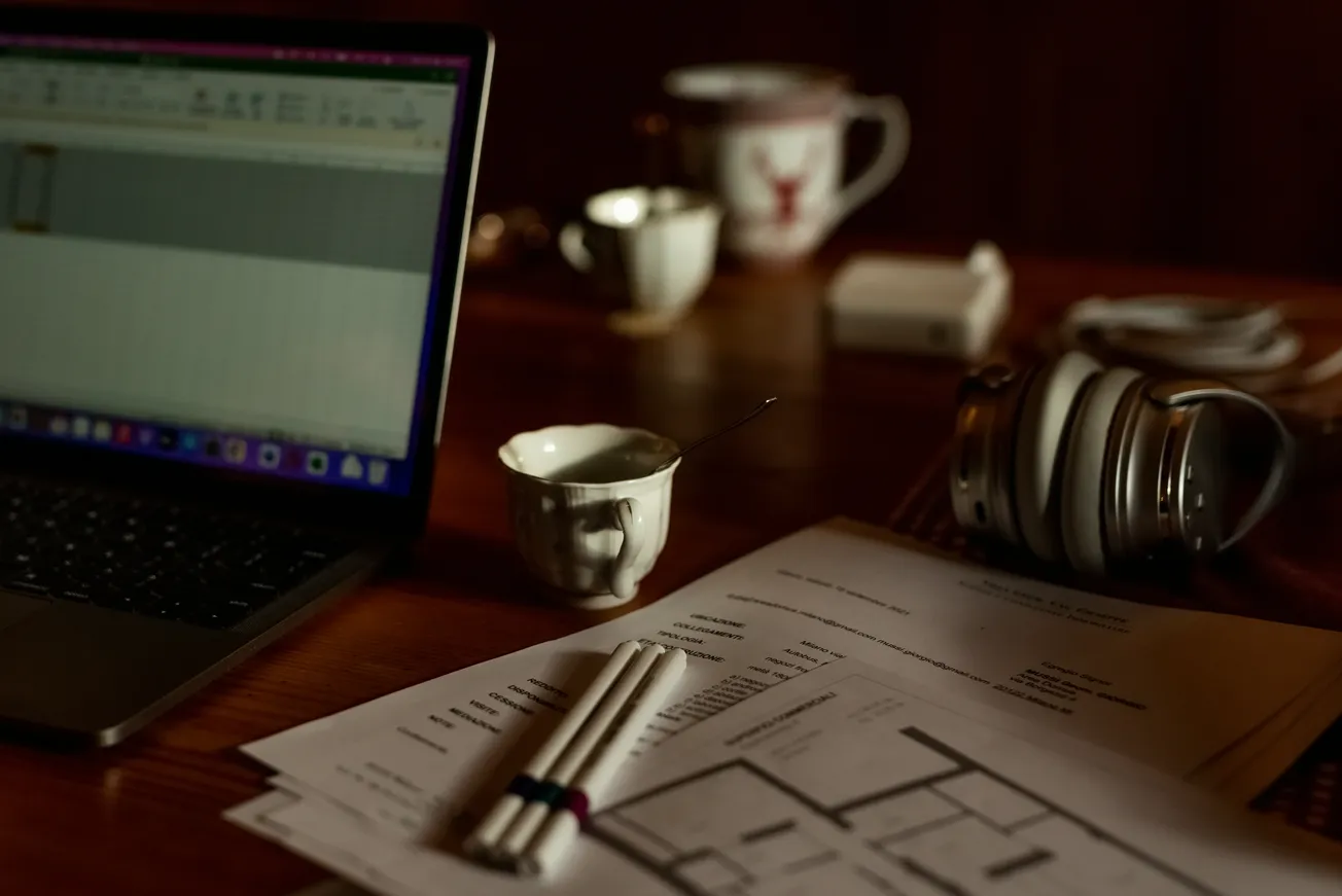 A dimly lit desk features a laptop showing a spreadsheet, architectural plans, pens, a cup with a spoon, headphones, and a mug, conveying a focused work atmosphere.
