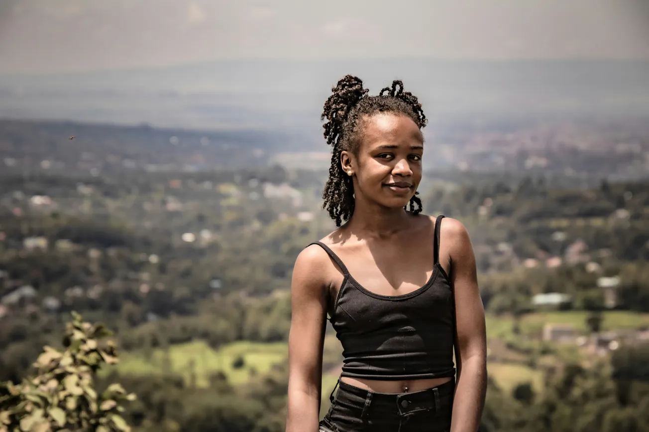 A young woman in a black top stands confidently against a scenic, blurred landscape of green fields and distant hills under a hazy sky.