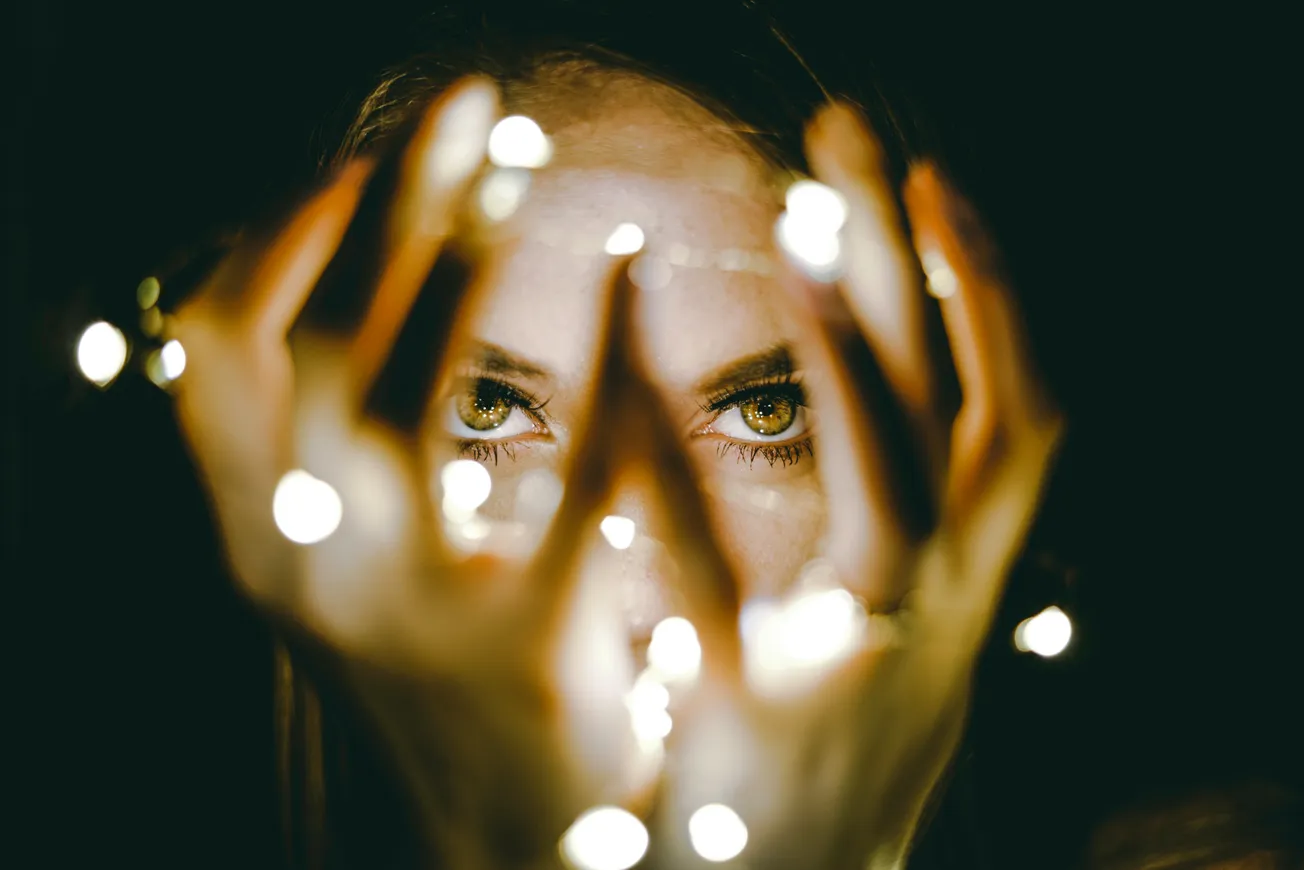 A person's intense eyes peer through their hands, which cradle glowing string lights in a dark setting, creating a mysterious and enchanting atmosphere.