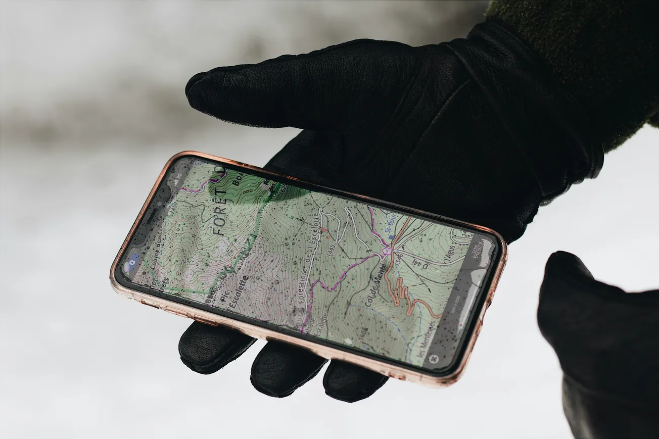 A gloved hand holds a smartphone displaying a topographic map, set against a snowy background. The scene conveys an outdoor adventure theme.