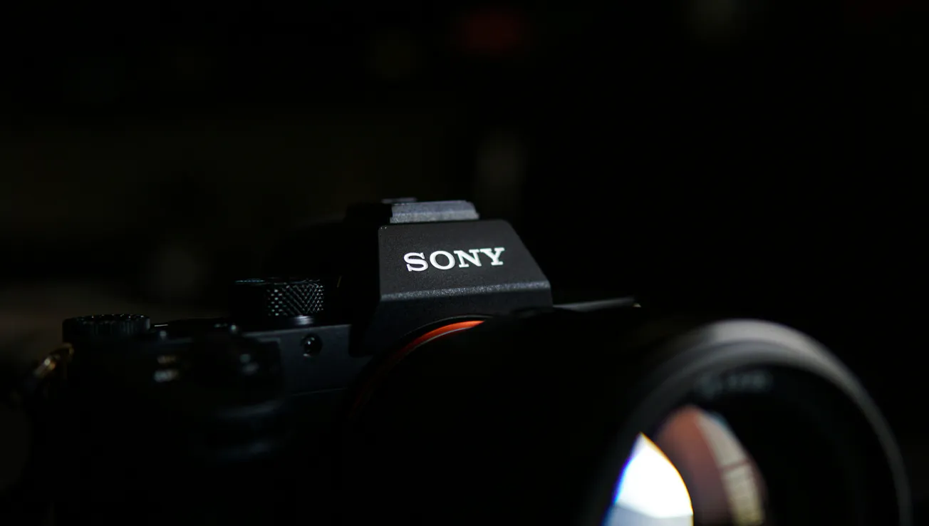 Close-up of a Sony camera in a dark setting, highlighting the brand logo. The lens reflects a soft light, creating a focused and professional tone.