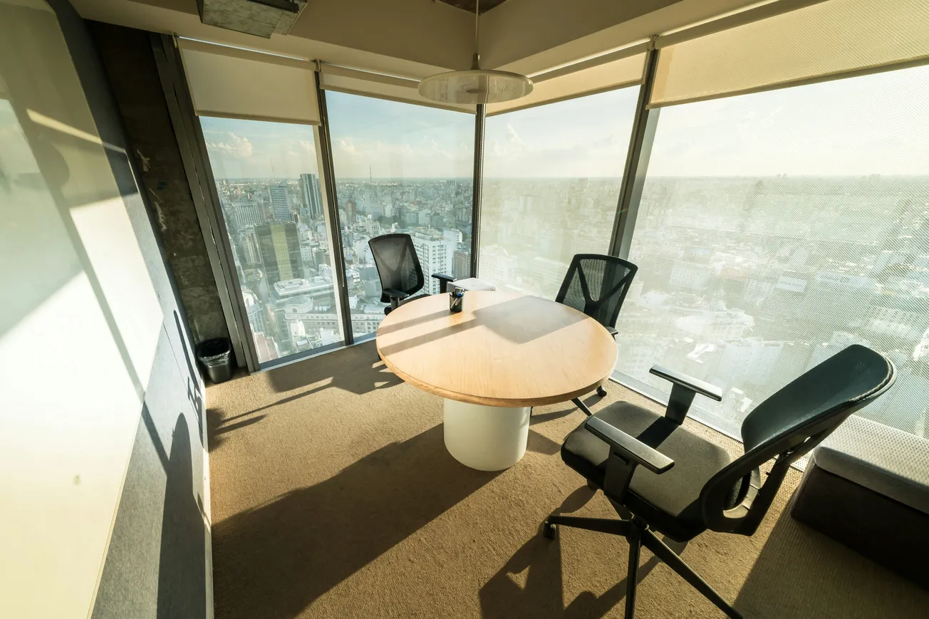 Bright office space with a round table and three modern chairs, surrounded by floor-to-ceiling windows offering a panoramic city view.
