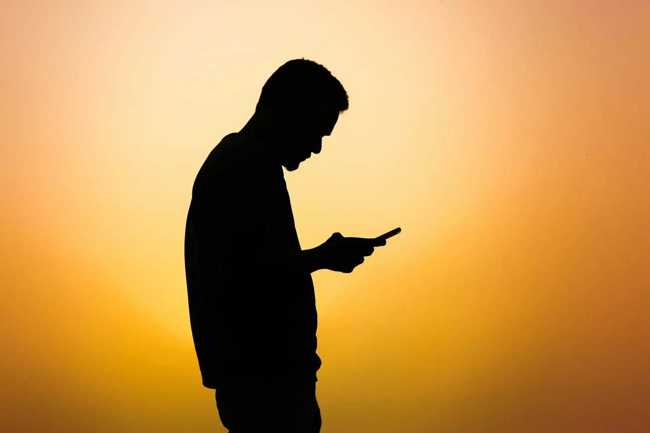 Silhouette of a person using a smartphone against a vibrant orange-yellow sunset, conveying a calm and reflective mood.