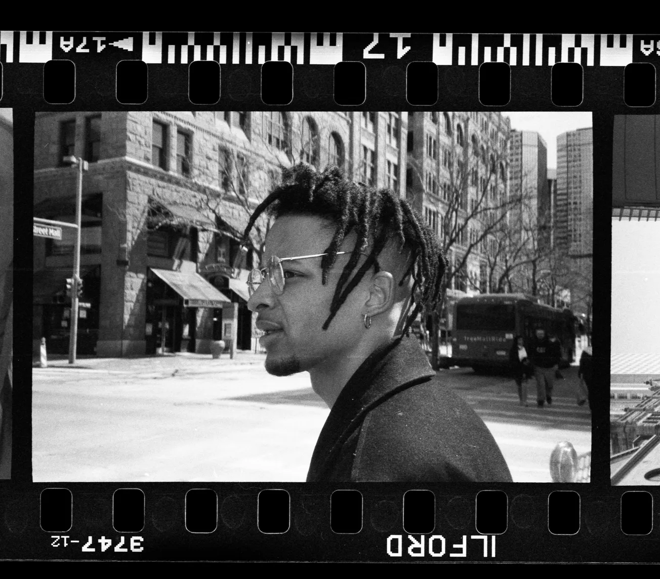 Black and white photo of a man with dreadlocks and glasses walking on a city street. Tall buildings and a bus are in the background, creating an urban vibe.