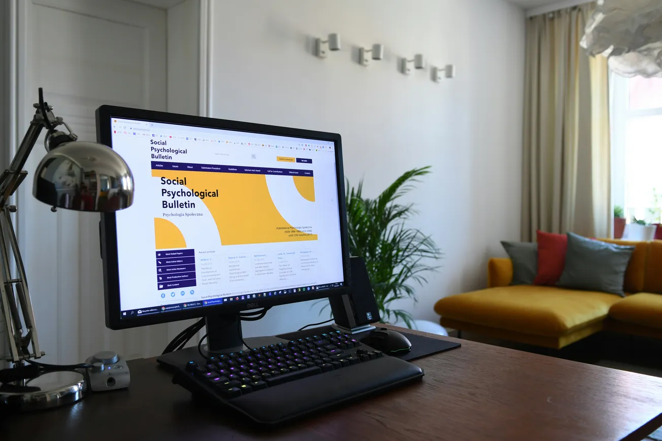 A home office with a monitor displaying a "Social Psychological Bulletin" website. The room has a modern desk lamp, plants, and a cozy yellow couch.