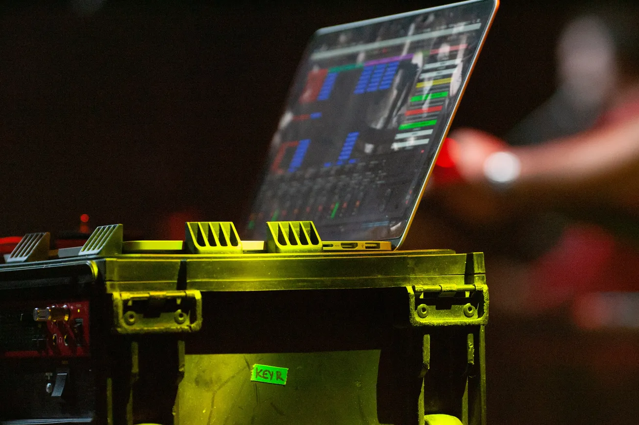 A laptop displaying colorful audio software on its screen sits atop a rugged equipment case on stage, with a dimly lit background suggesting a concert setting.