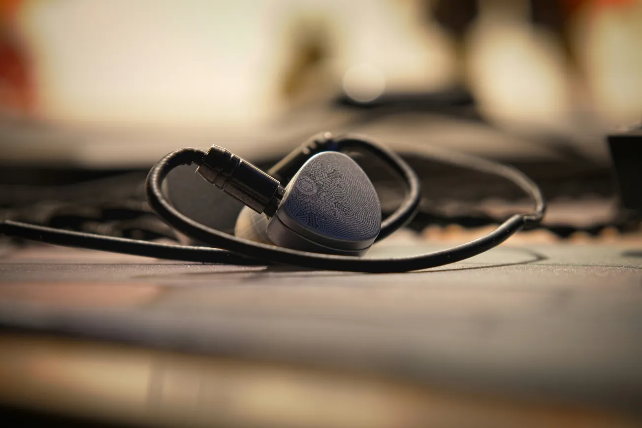 A pair of professional gray in-ear monitors with intricate etched patterns on the faceplates rest on a desk with their black cables looped.