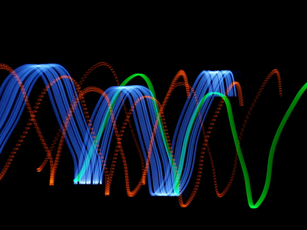 Abstract light painting with blue, orange, and green light trails forming overlapping wavy patterns against a black background, creating a dynamic and vibrant energy.
