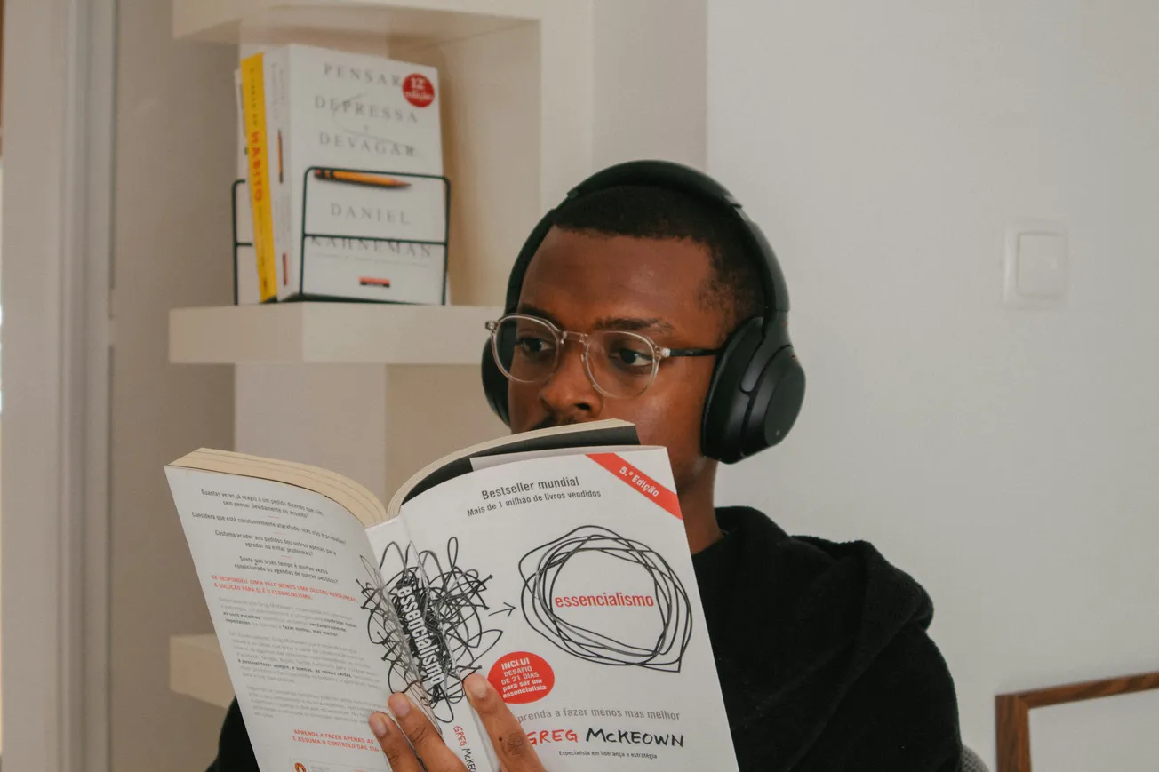 A person with glasses and headphones reads "Essentialism" by Greg McKeown in a cozy room. Books are neatly arranged on a white shelf behind.