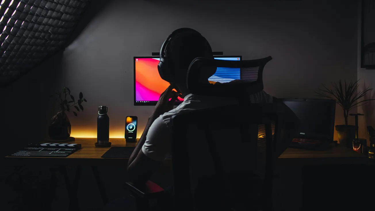 A person in silhouette sits at a dark, moody computer desk with dual screens. Warm lighting highlights a plant, water bottle, and open laptop.