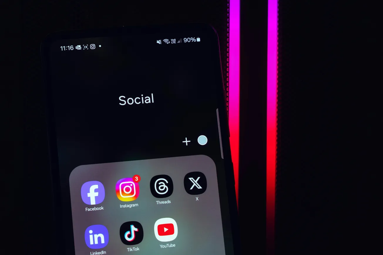 Smartphone screen displaying social media apps like Facebook, Instagram, Threads, X, LinkedIn, TikTok, and YouTube on a dark background.