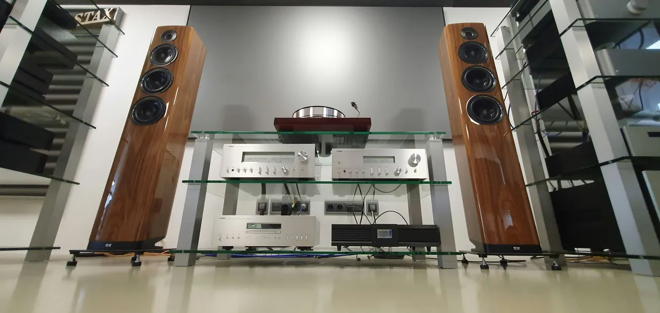 A high-end audio setup with wooden floor-standing speakers on either side; three glass shelves hold silver amplifiers and audio equipment, conveying a modern, sophisticated tone.