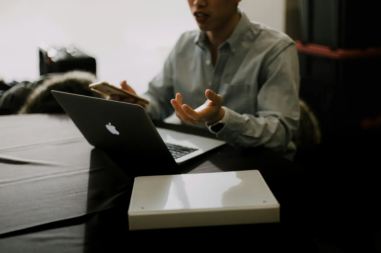 A person in a denim shirt gestures expressively while holding a phone and sitting at a table with a laptop. The setting appears informal and focused.