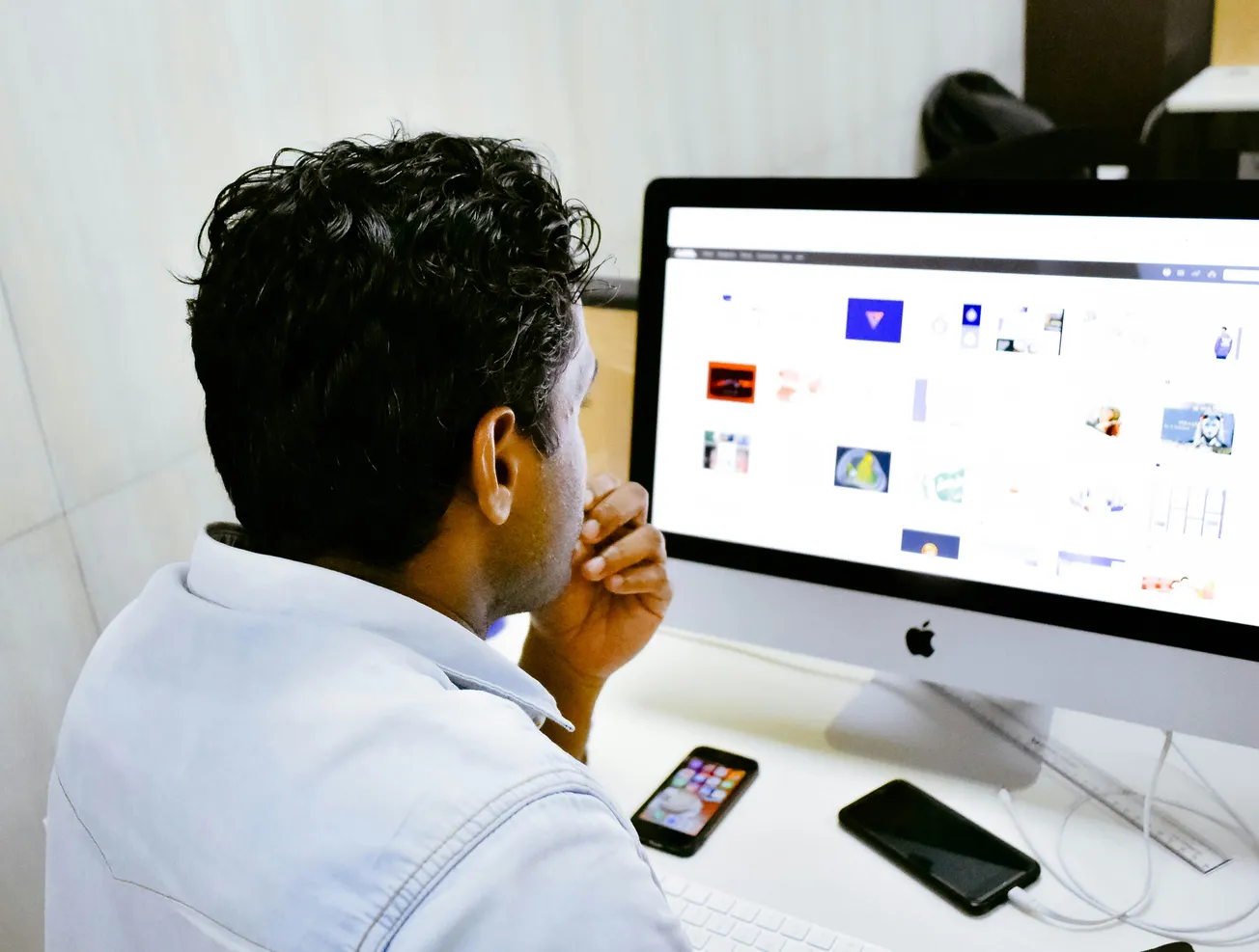 A man, deep in thought, focuses on a desktop computer displaying various images. A smartphone and tablet lie on the white desk, suggesting multitasking.