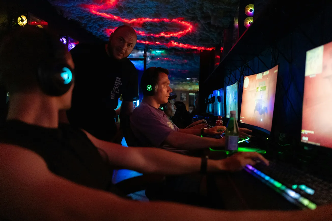 In a dimly lit gaming room, three people wearing headphones focus intently on their glowing screens. The ceiling displays vivid red and blue lights, adding a vibrant atmosphere.