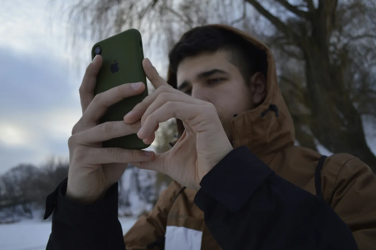 A person in a brown jacket stands outdoors on a snowy day, focused on a smartphone. Bare trees and a cloudy sky form the background, suggesting winter.