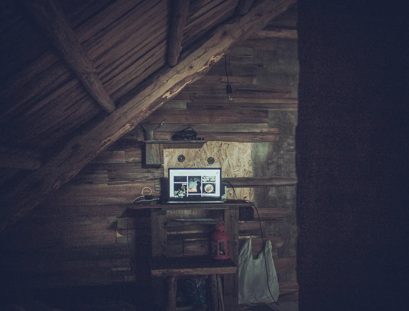 A dimly lit rustic attic with a wooden desk under a slanted roof. A laptop displaying images sits on the desk, next to a red lantern and a hanging bag. The atmosphere is cozy and secluded.
