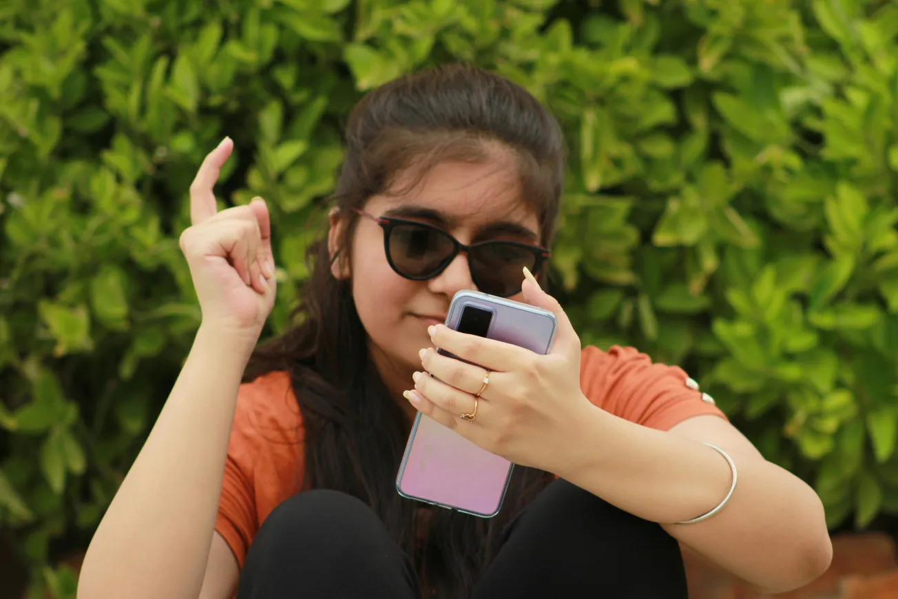 Young woman in sunglasses holds a smartphone, smiling as she gestures with one hand. She's seated against lush green foliage, wearing a casual orange top.